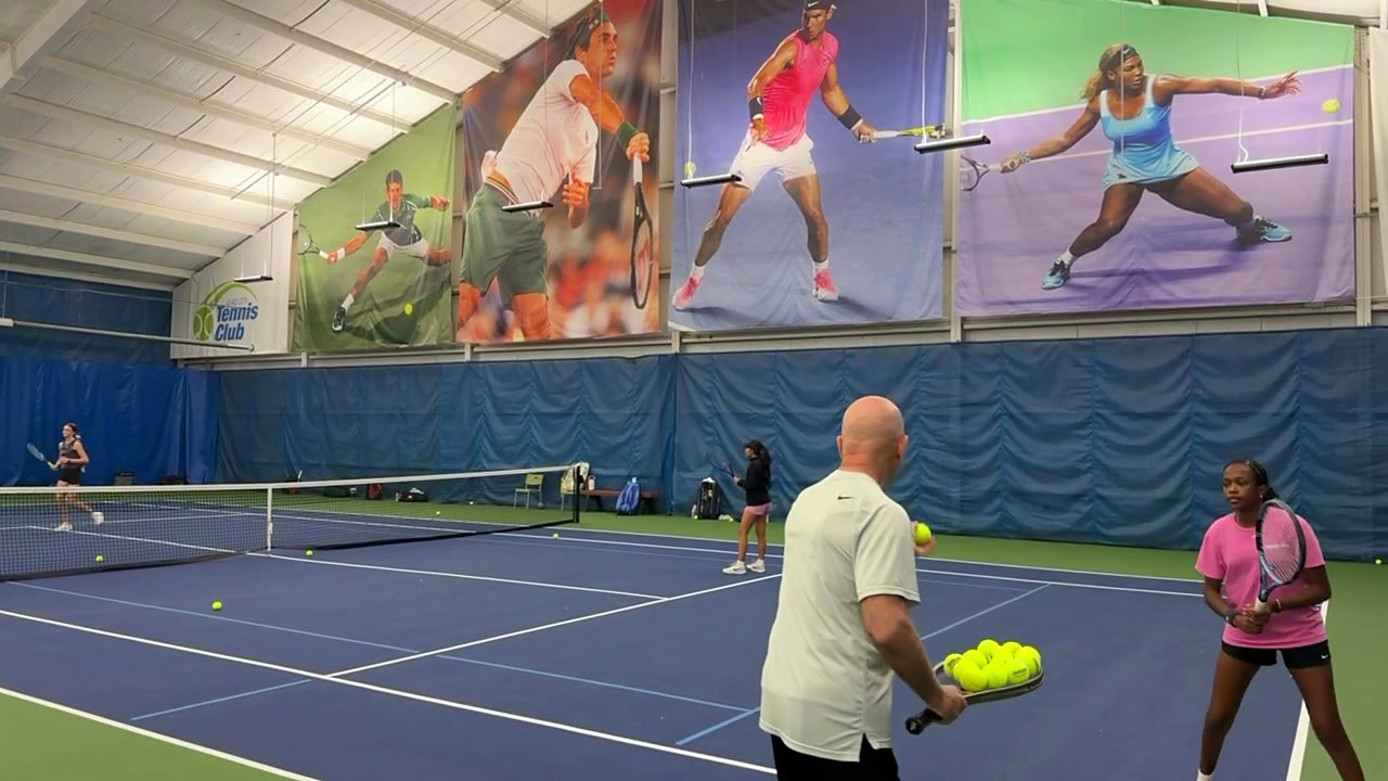 A coach feeding balls to a junior player on the indoor courts