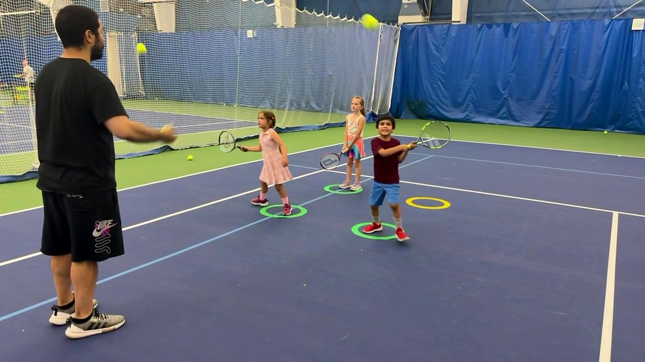 A coach tossing balls to young players during a beginner tennis class